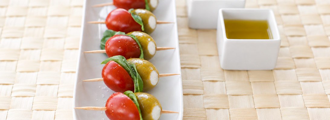 Fuente blanca con pinchos de tomate cherry albahaca y aceitunas grandes rellenas de queso junto a cuencos de aceite y vinagre Fuente blanca con pinchos de tomate cherry albahaca y aceitunas grandes rellenas de queso junto a cuencos de aceite y vinagre